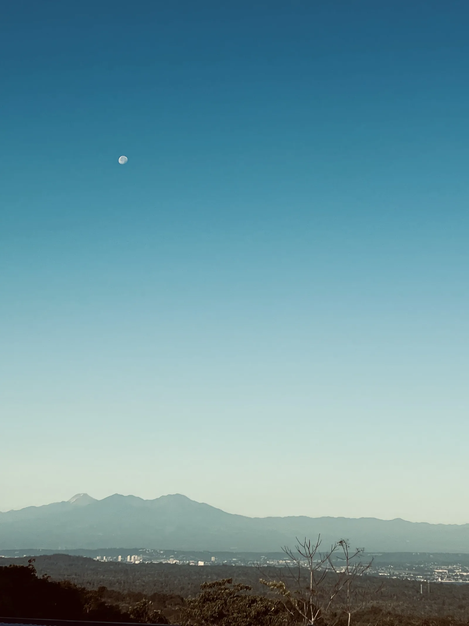 A clear blue sky with a daytime moon above distant mountains and the buildings of Davao City.