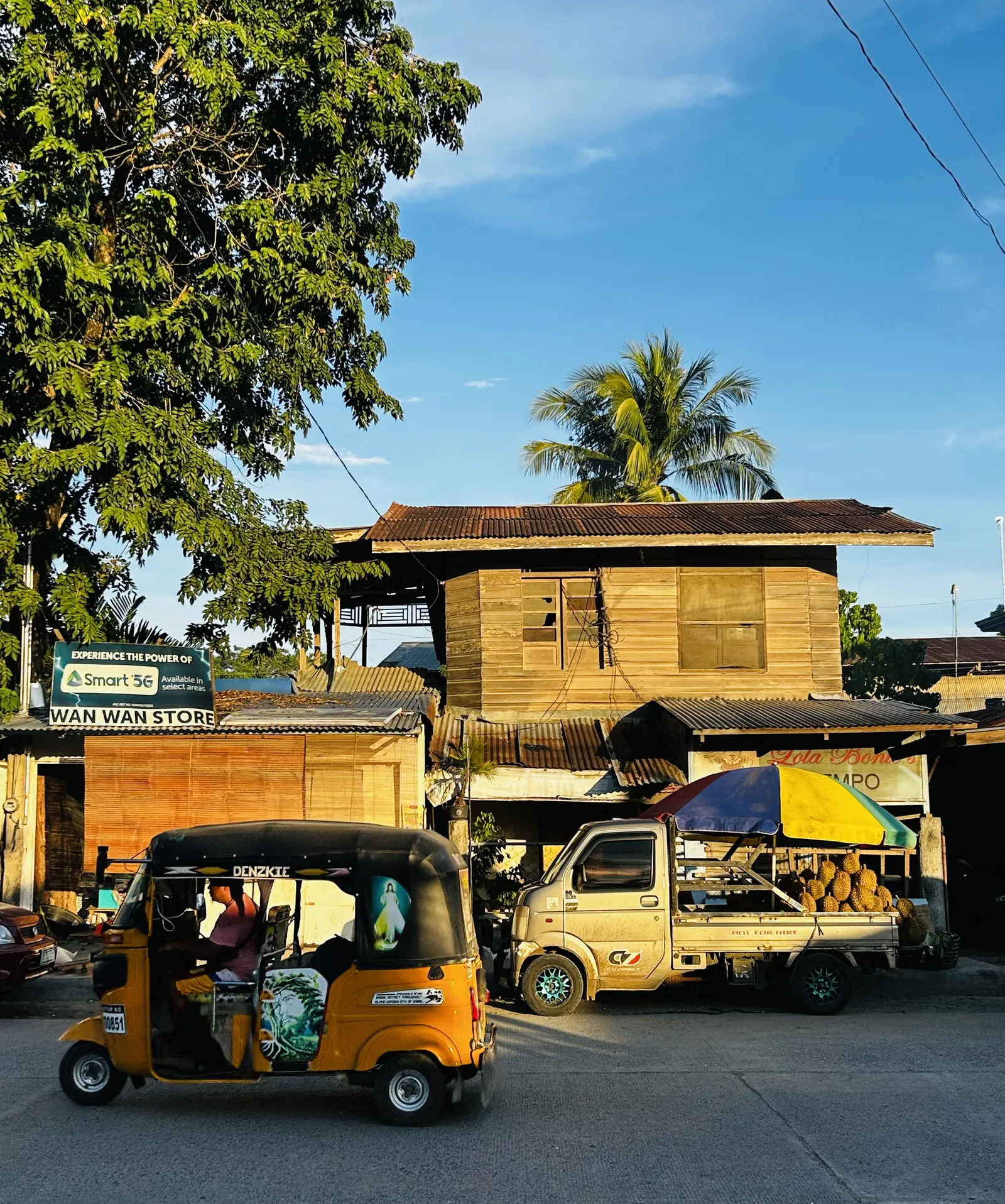 A street on Samal in late afternoon light, with a yellow tricycle, a small truck loaded with durian, and a wooden two-storey building.